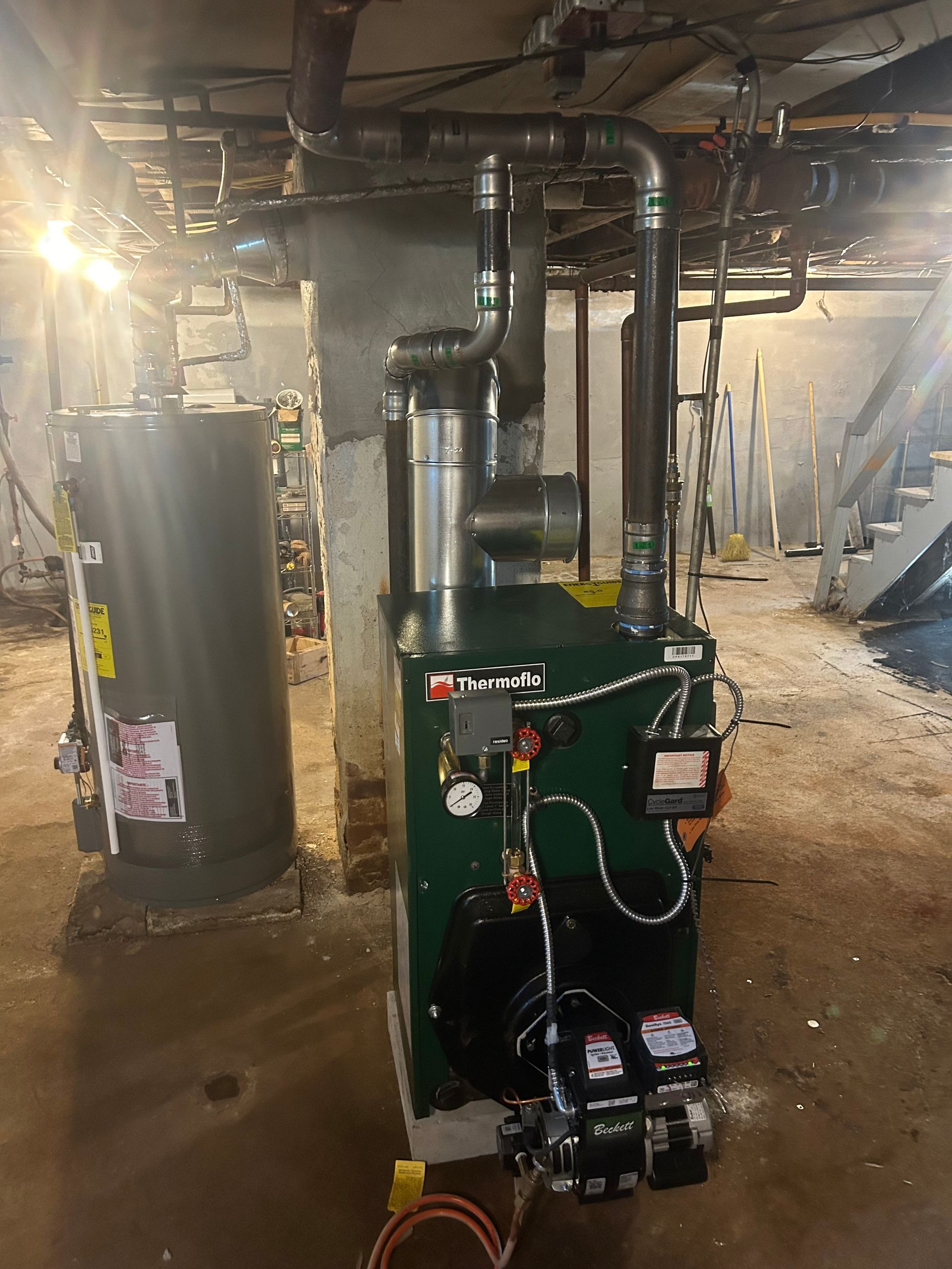 A green oil-fired boiler and a grey hot water heater stand in a basement with exposed ceiling pipes and concrete floors.