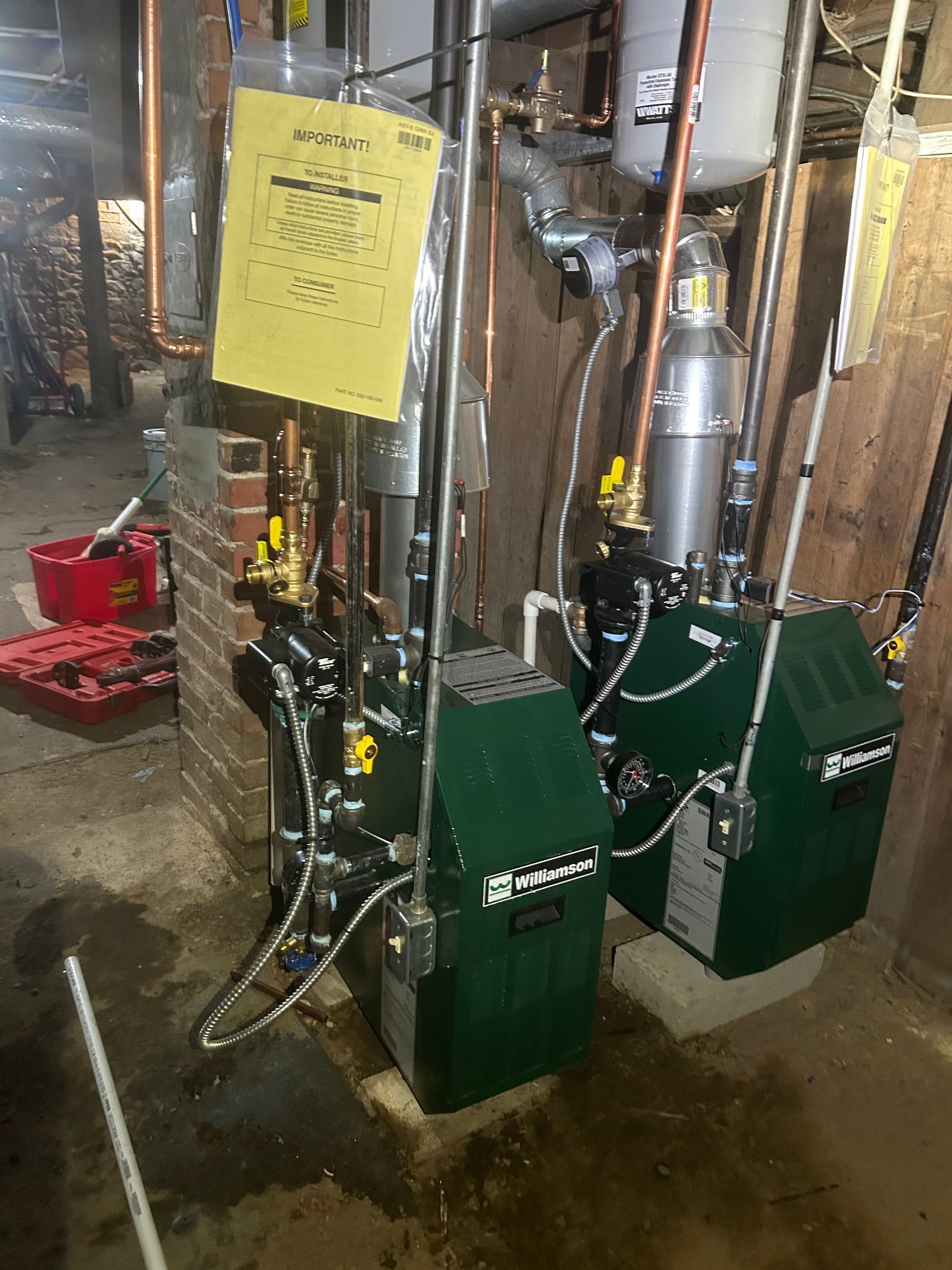 Two green Weil-McLain boilers installed side-by-side in a basement, with copper piping and a yellow sign attached.