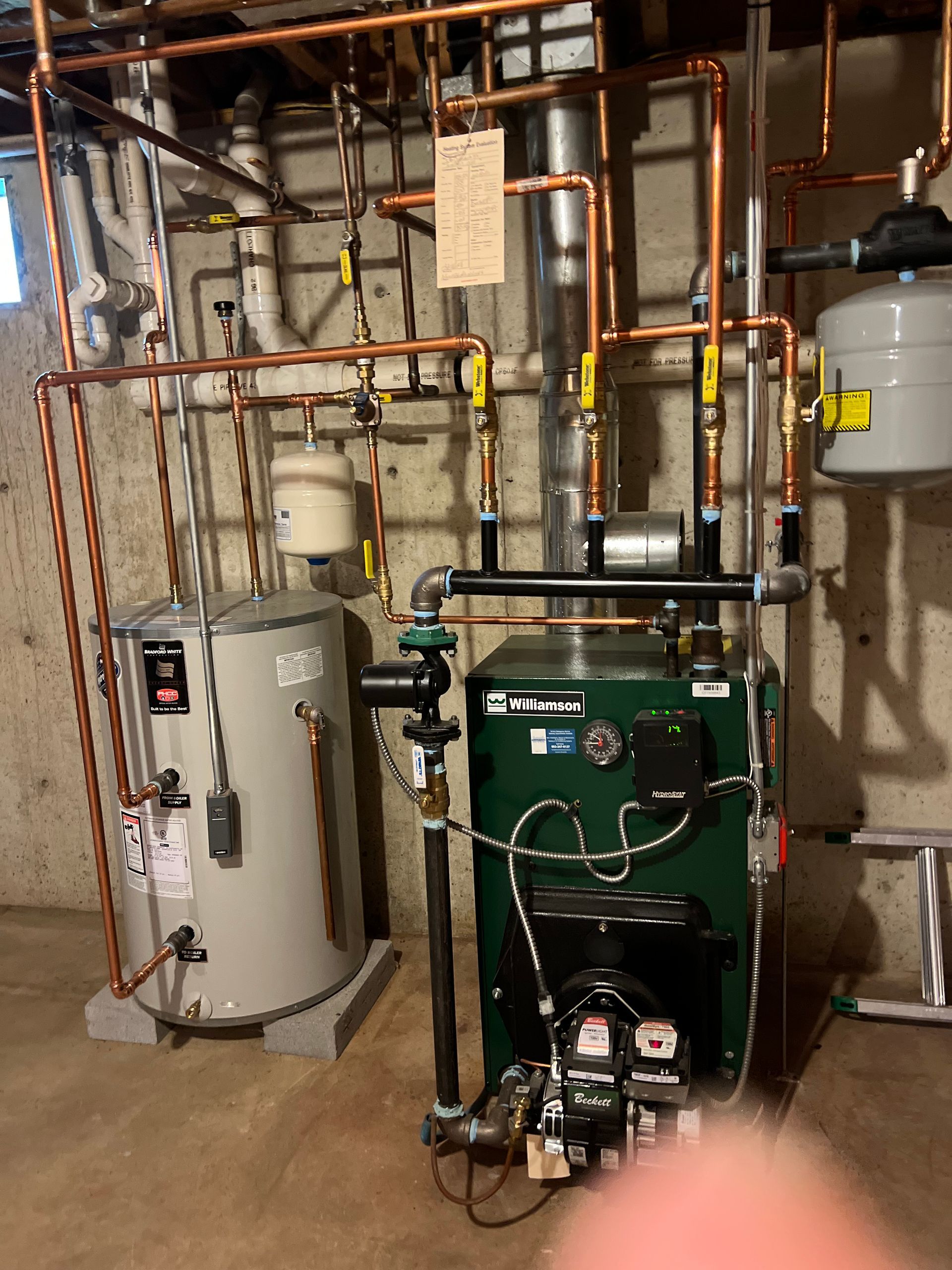 A basement mechanical room showing a green heating boiler, a grey water tank, and extensive copper piping connections.