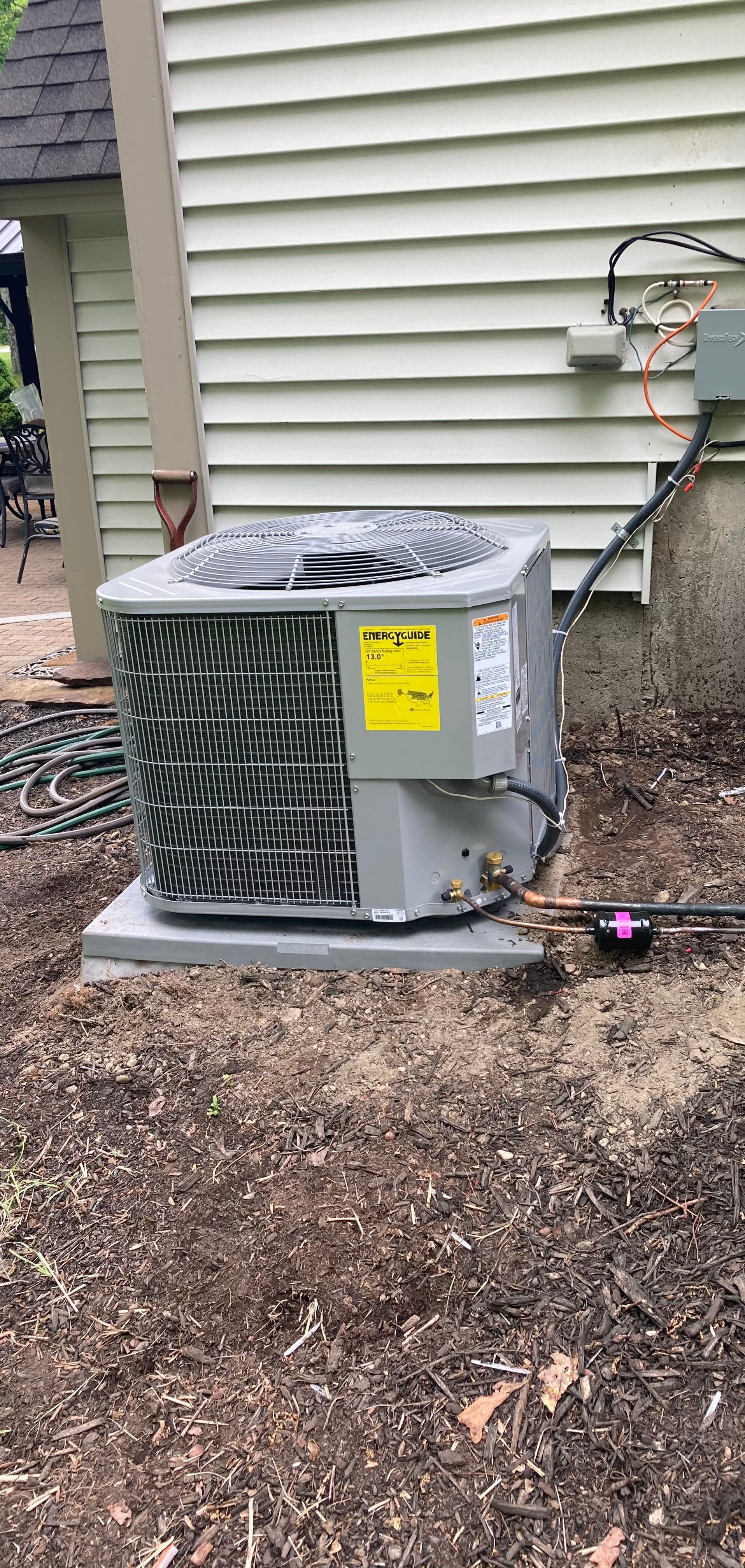 A gray central air conditioning unit sits on a concrete pad next to the vinyl-sided wall of a house.