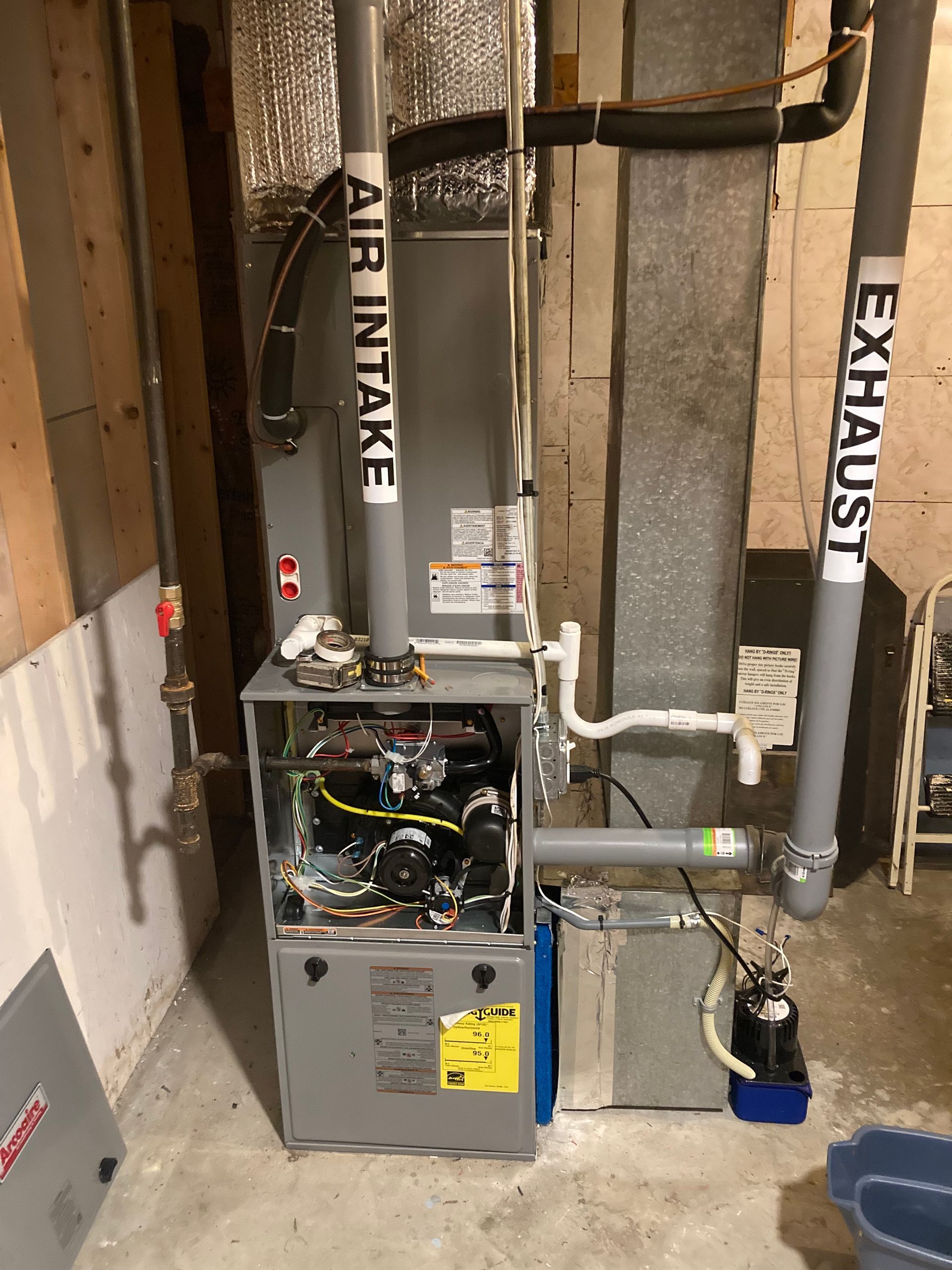 A high-efficiency gas furnace stands in a basement with labeled PVC air intake and exhaust pipes connected to the unit.