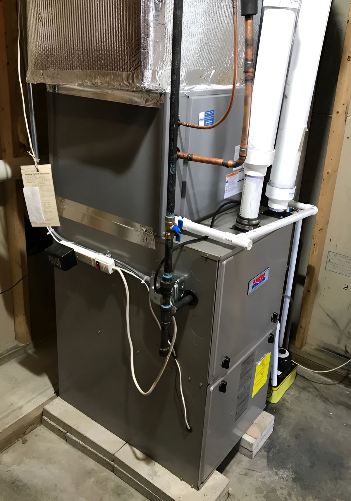 A grey HVAC furnace unit sits on a concrete floor in a basement, featuring vertical white pipes and copper tubing.