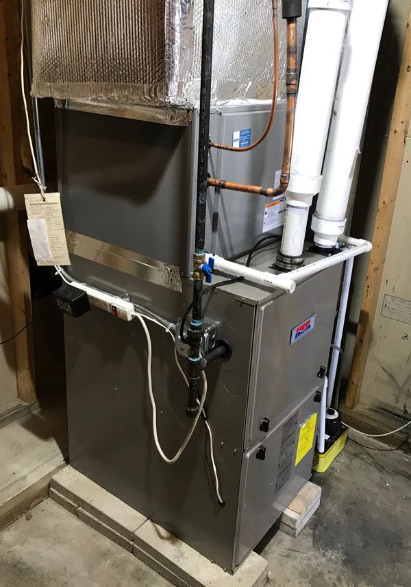 A gray HVAC furnace unit with white PVC intake and exhaust pipes, standing on a concrete floor in a basement.