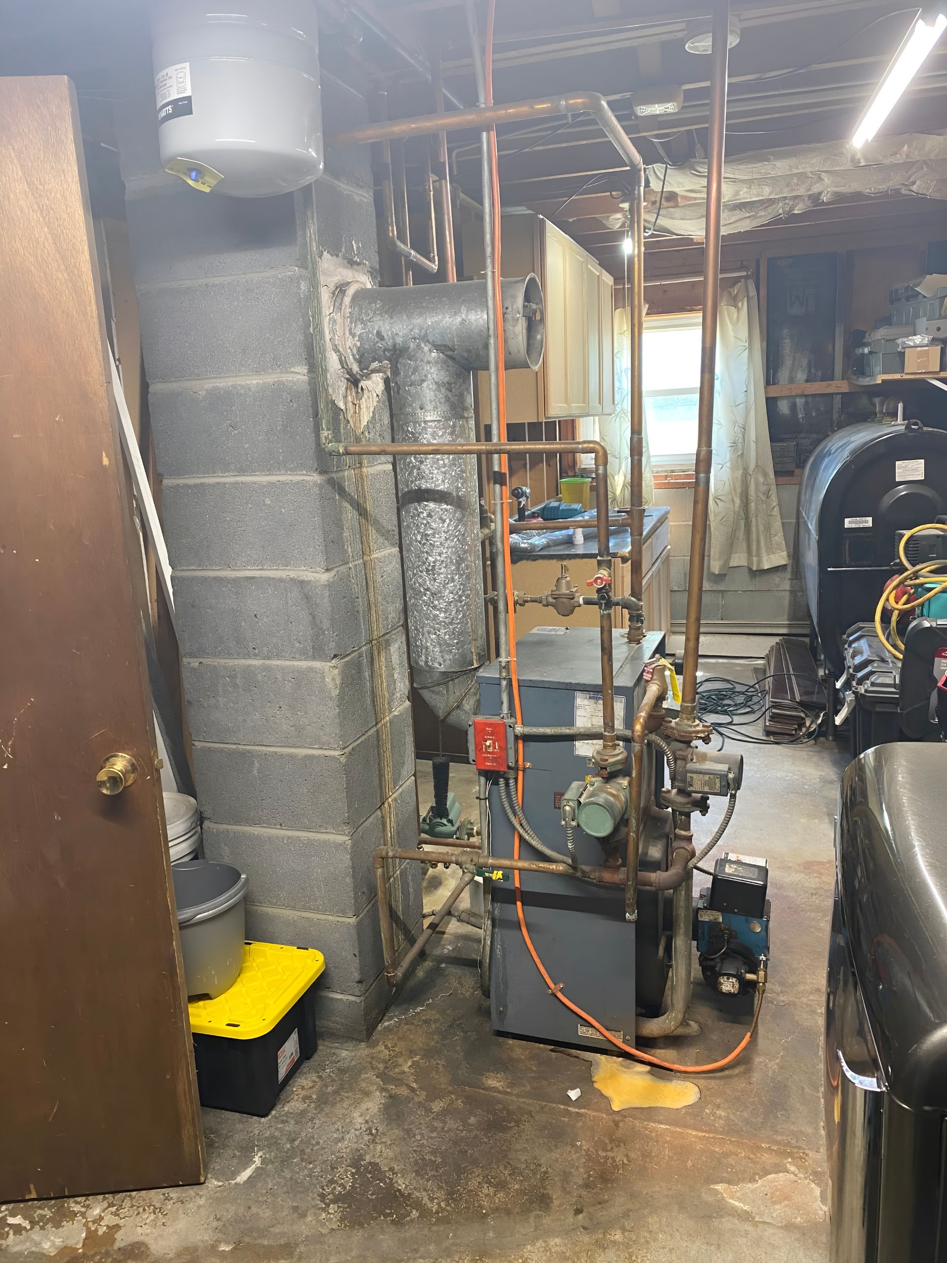 A utility basement featuring a grey boiler system, copper piping, a vent pipe, and an expansion tank on a concrete wall.
