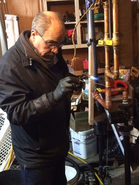 A person in a black jacket and gloves uses a blowtorch to solder copper pipes in a workshop.