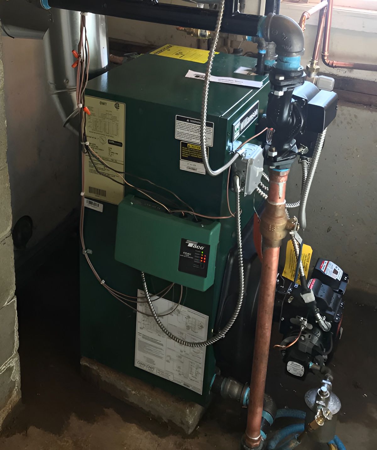 A green residential boiler unit with copper pipes and a control panel, installed in a basement.