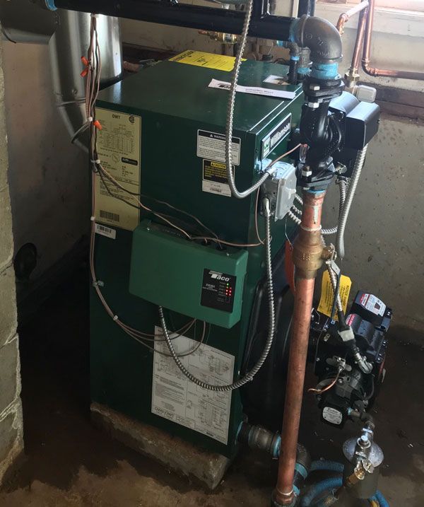 A dark green residential heating boiler with copper piping and electrical controls in a basement setting.