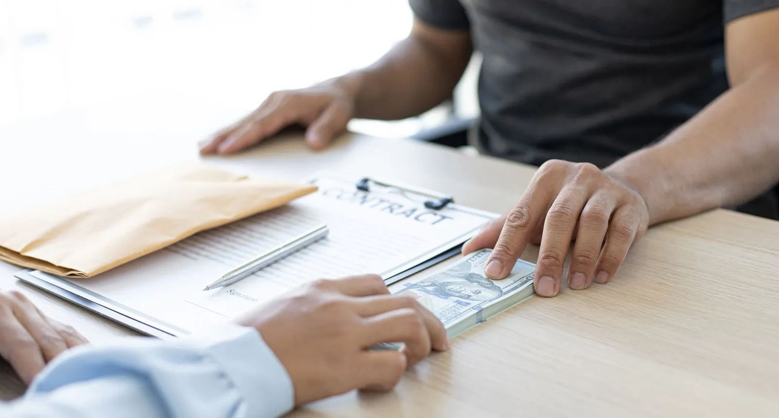 Person handing a wad of cash to another person over a contract on a desk, near an envelope.