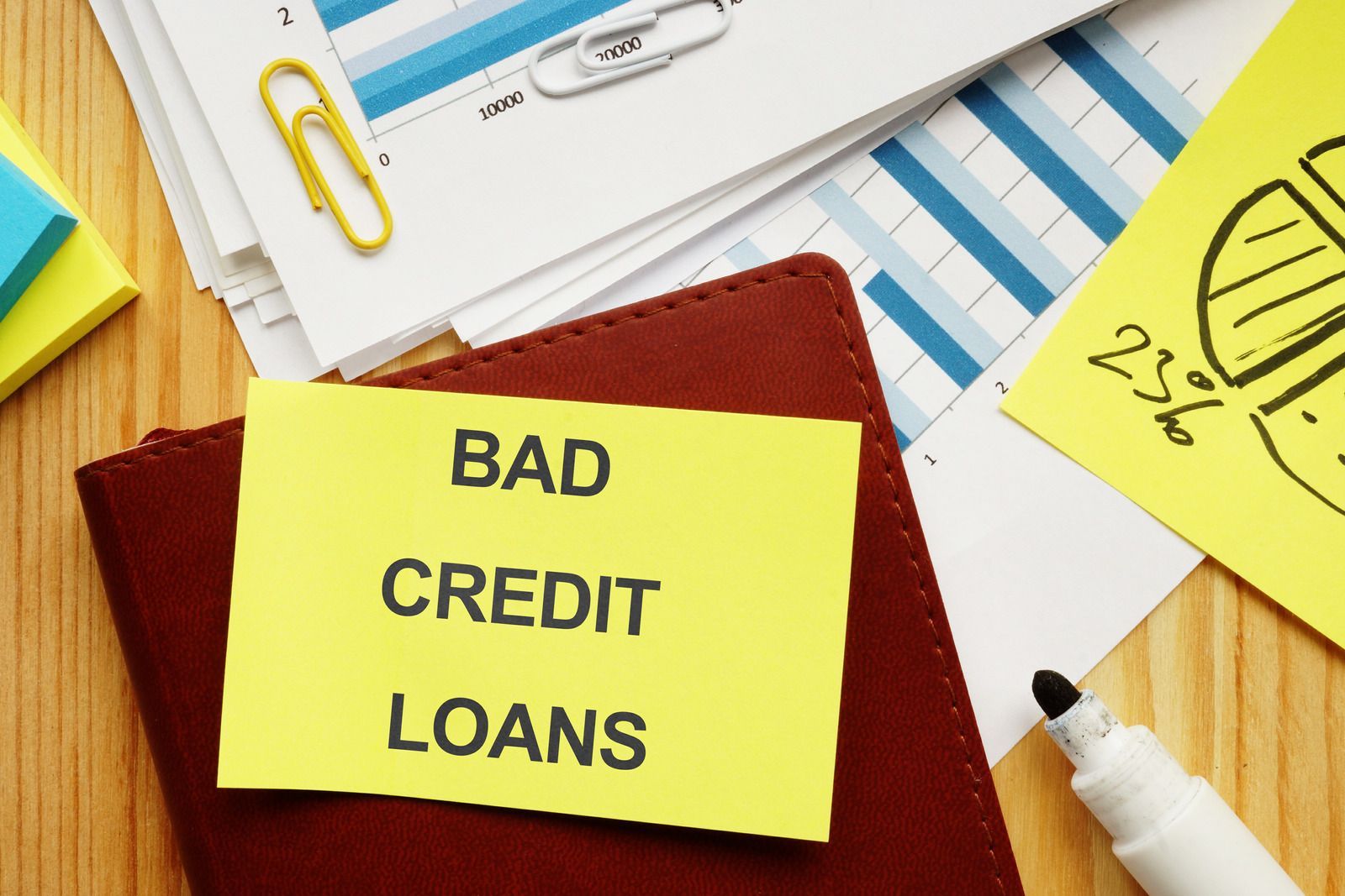 Yellow note reading “BAD CREDIT LOANS” on a desk with papers, pen, and paperclips