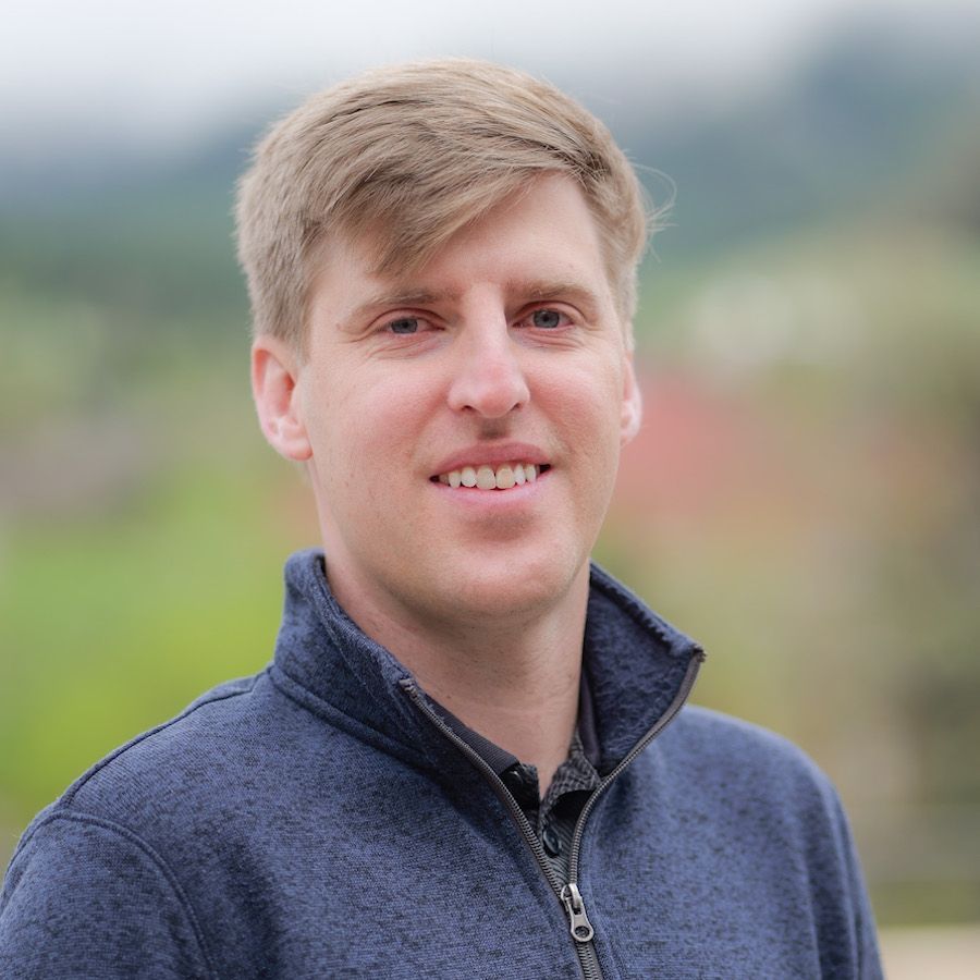 Man with light hair and blue sweater smiling outside.