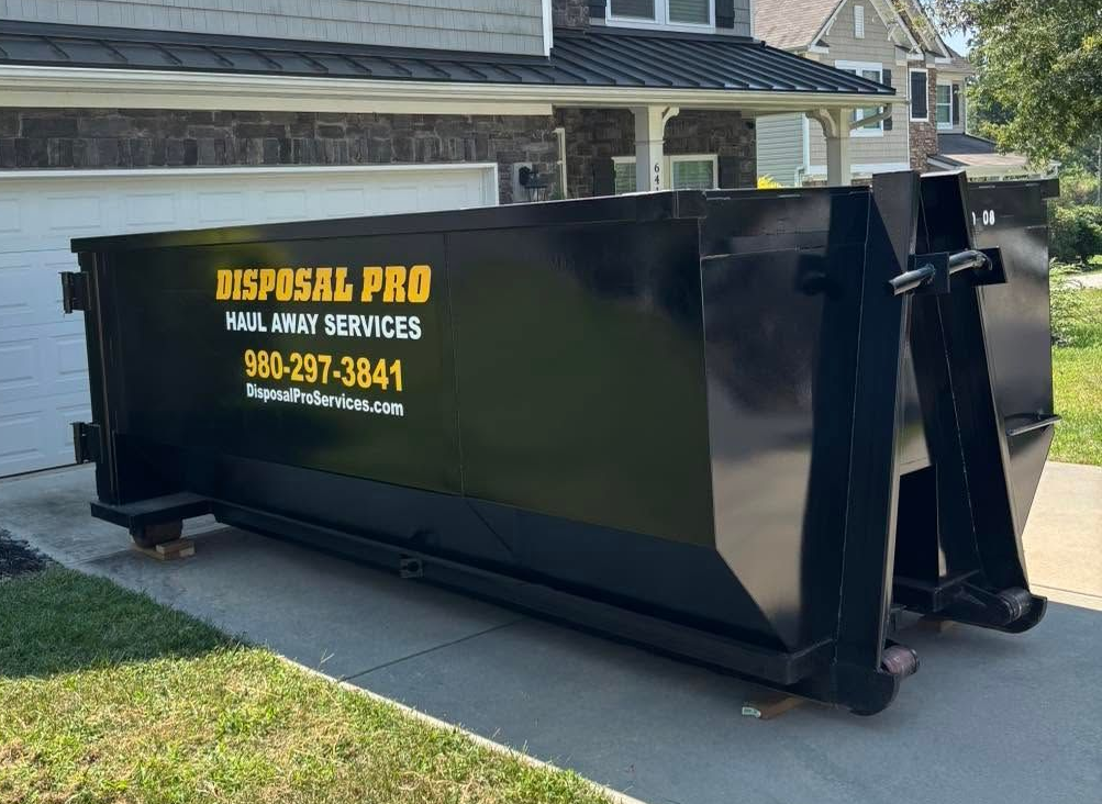 Black dumpster container on a driveway in front of a house with “NISPASAL PRO” signage.
