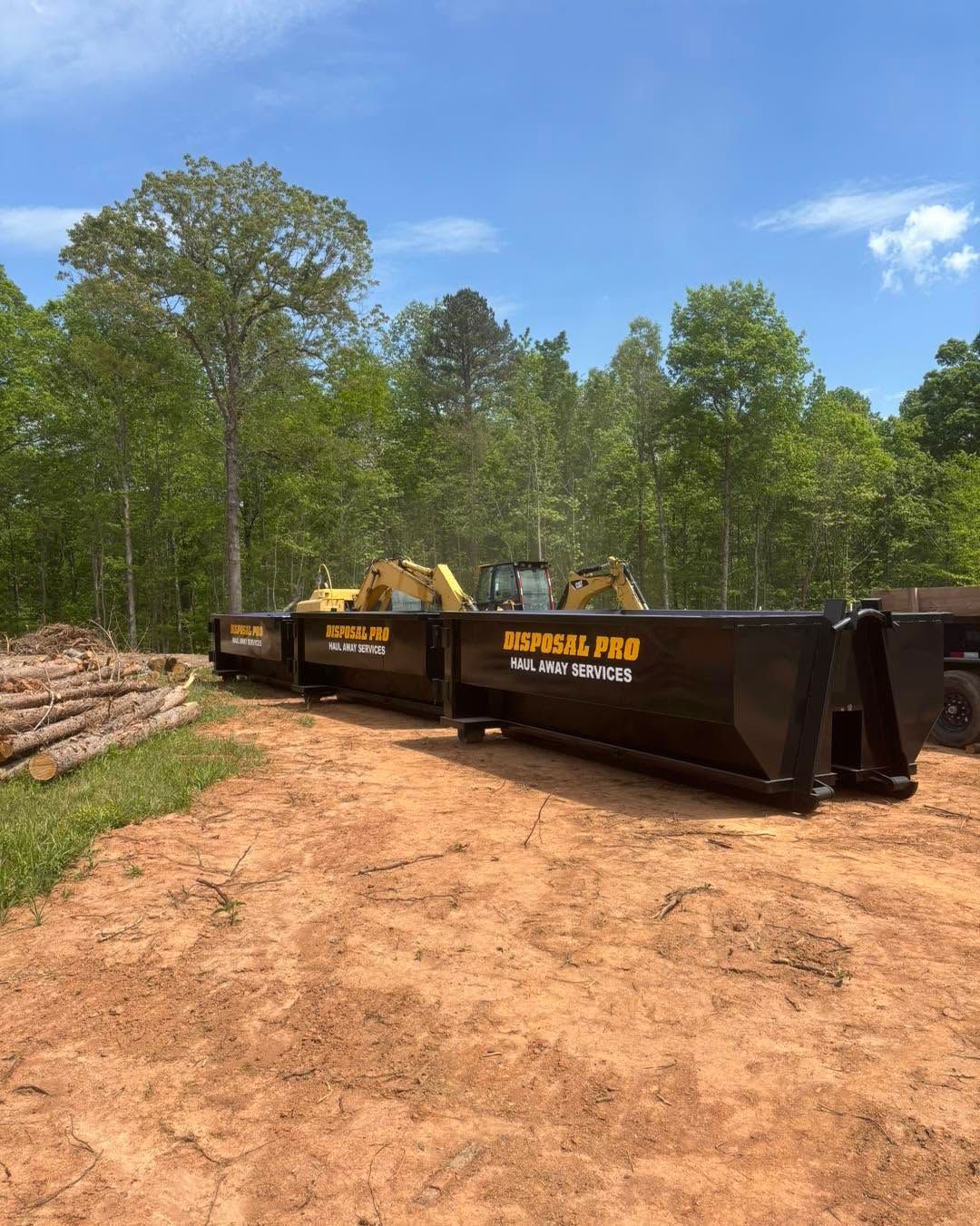 Yellow excavators beside a black dumpster on a dirt clearing with stacked logs and trees behind