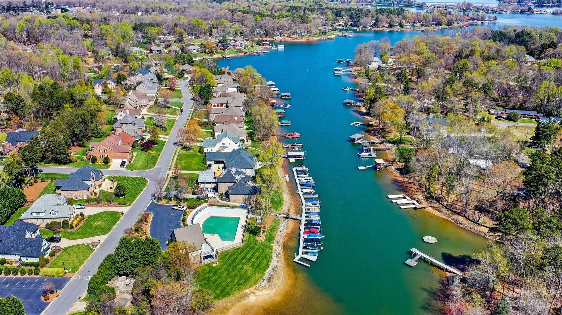 Aerial view of a narrow blue canal lined with houses, docks, boats, and spring-green trees.