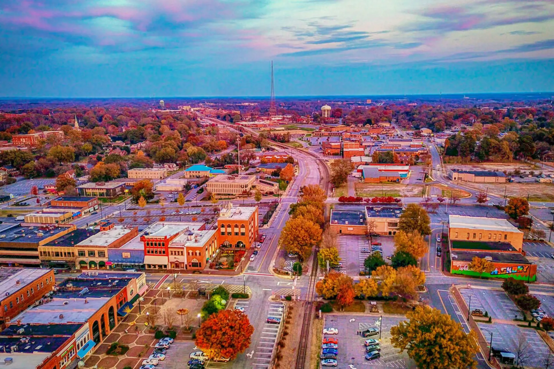 Aerial view of a small town at sunset with tree-lined streets, buildings, and cars on a main road.