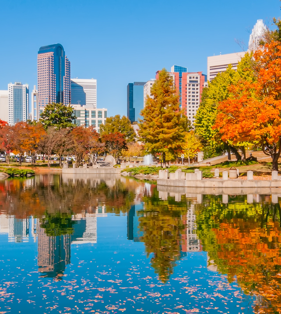 Autumn city skyline reflected in a calm lake, with colorful trees along the shore.