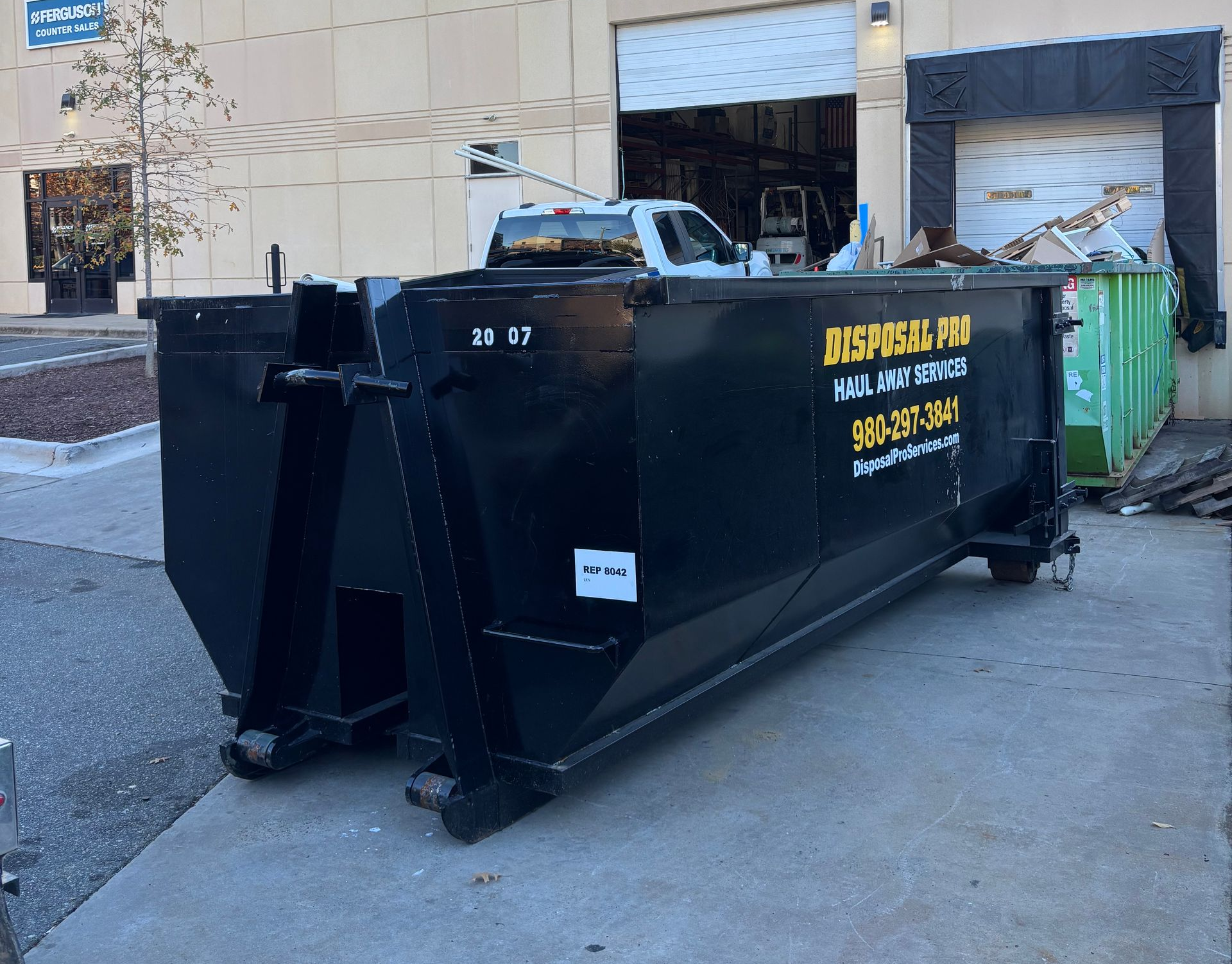 Large black construction dumpster on a sidewalk beside a building, with yellow lettering and green dumpsters behind it