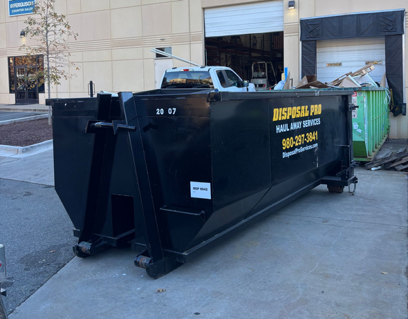 Large black dumpster with yellow lettering beside green recycling bins outside a warehouse loading dock