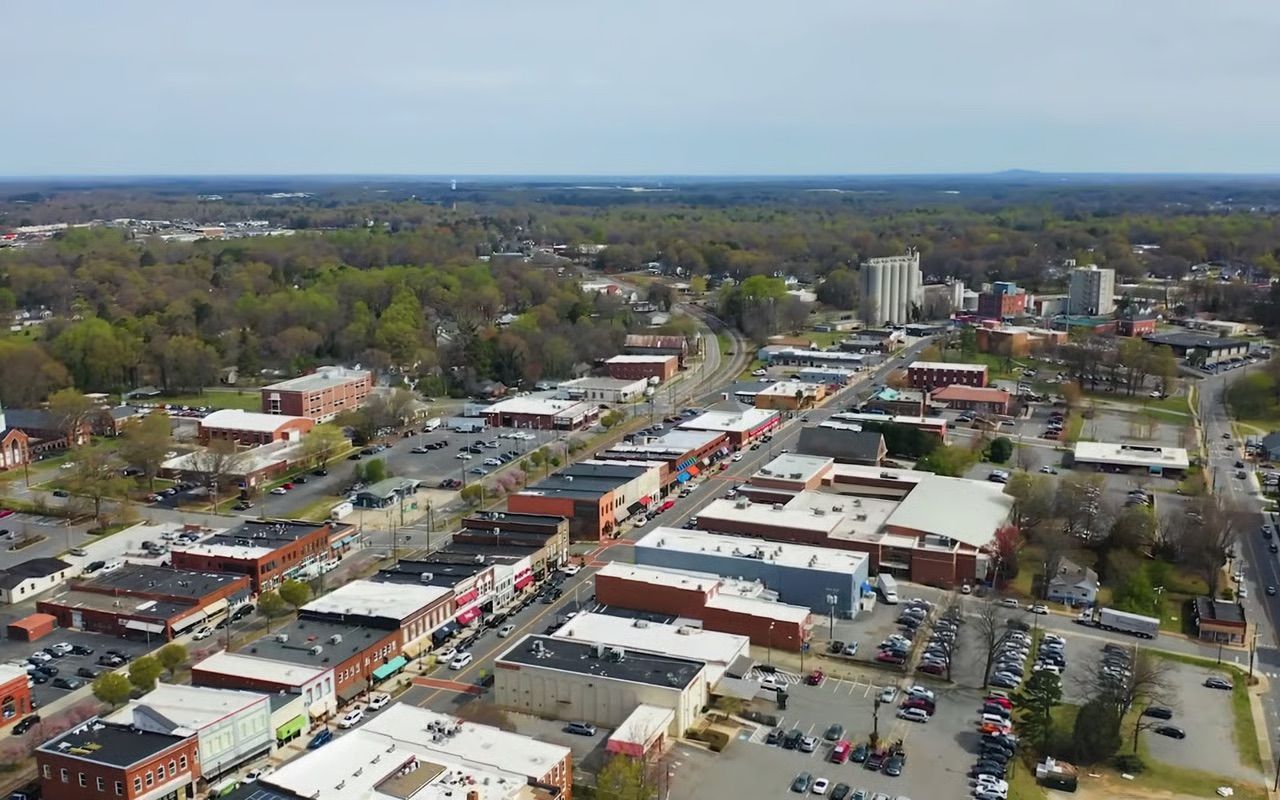 Aerial view of a small downtown with shops, parking lots, and tree-lined streets under a clear sky