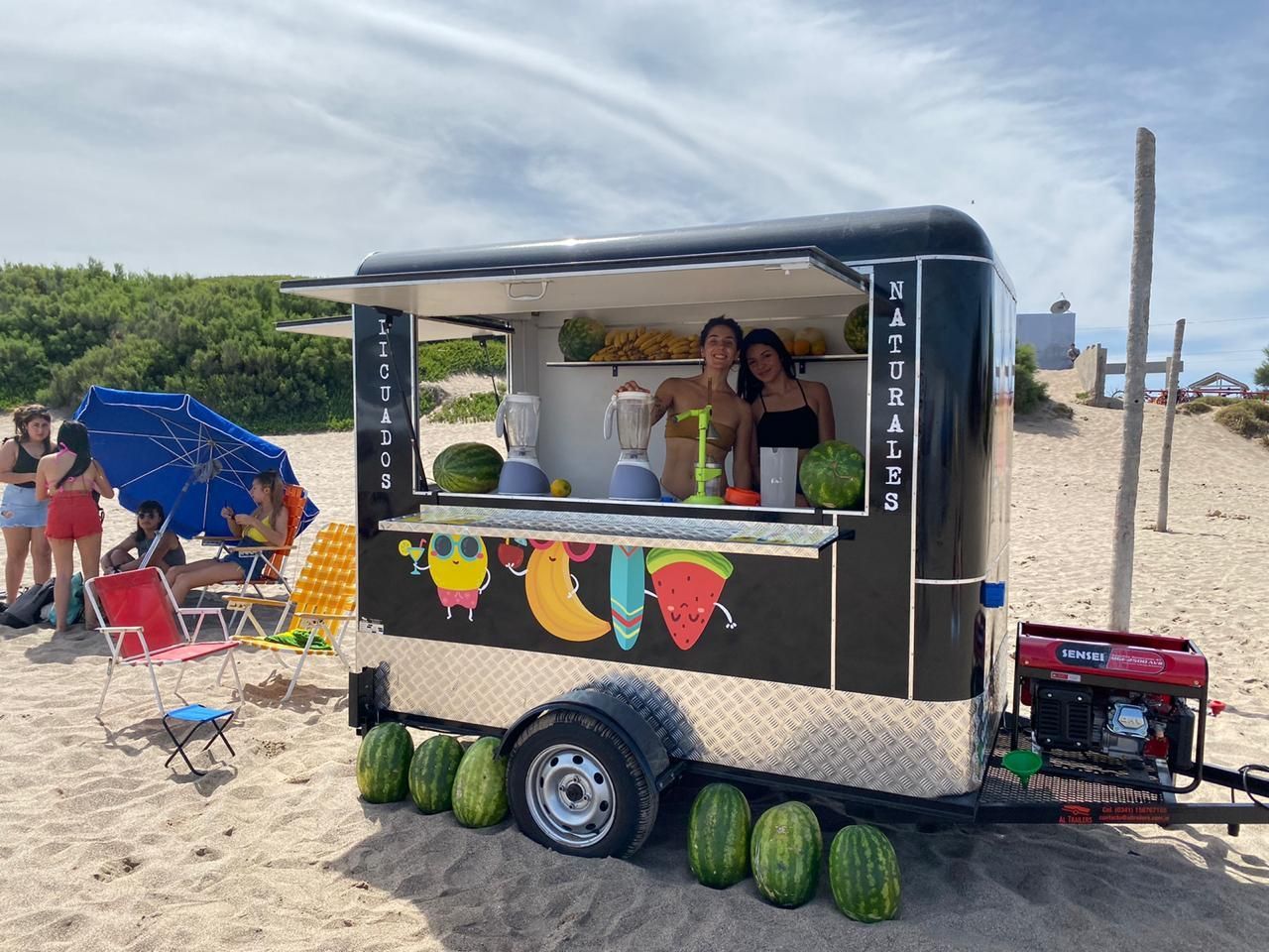 Un carrito de bebidas negro con una colorida decoración de frutas y personas sirviendo bebidas en la orilla arenosa.