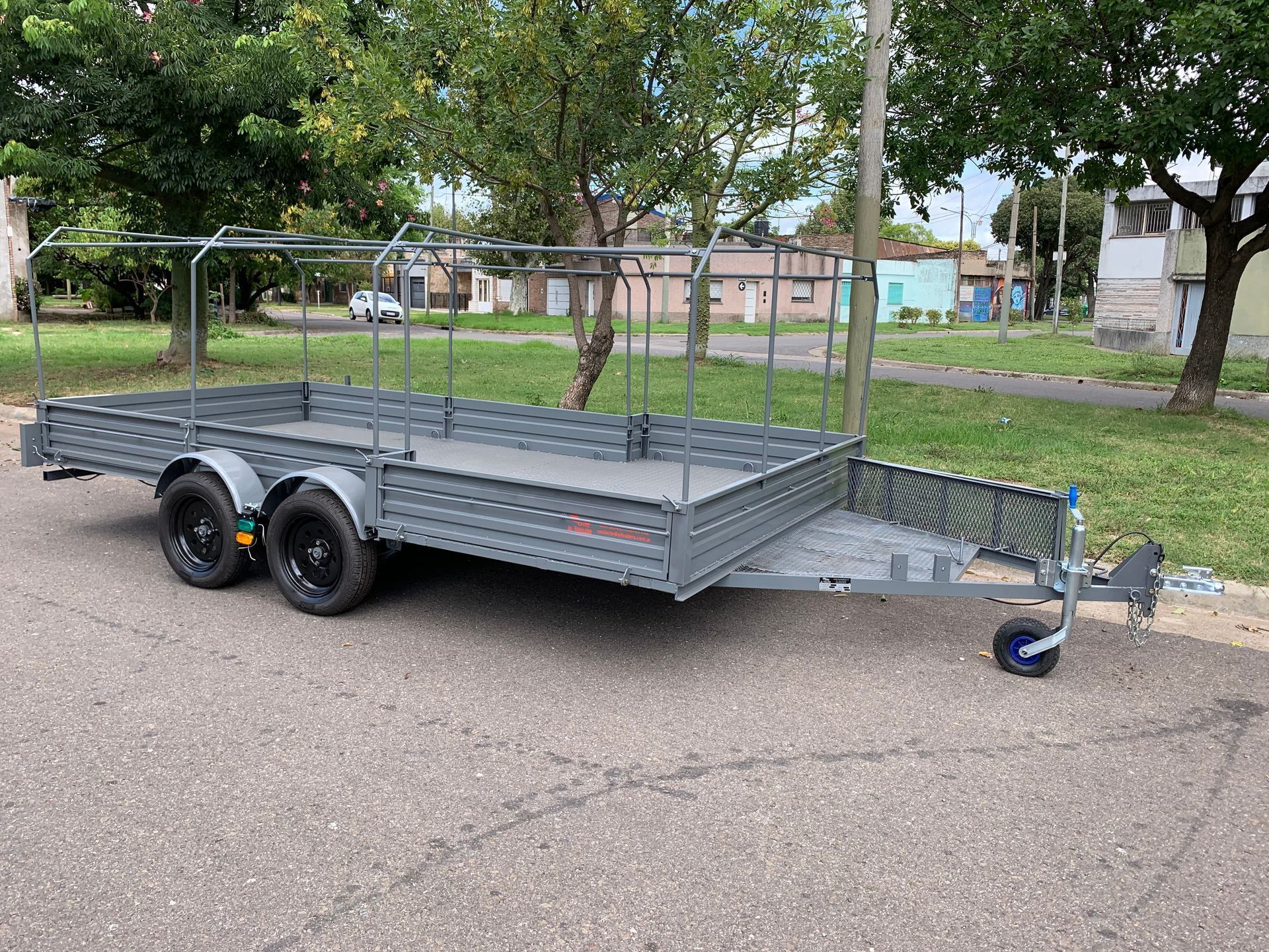 Remolque utilitario gris de doble eje con laterales de malla y enganche estacionado en el pavimento cerca de un terreno cubierto de césped.
