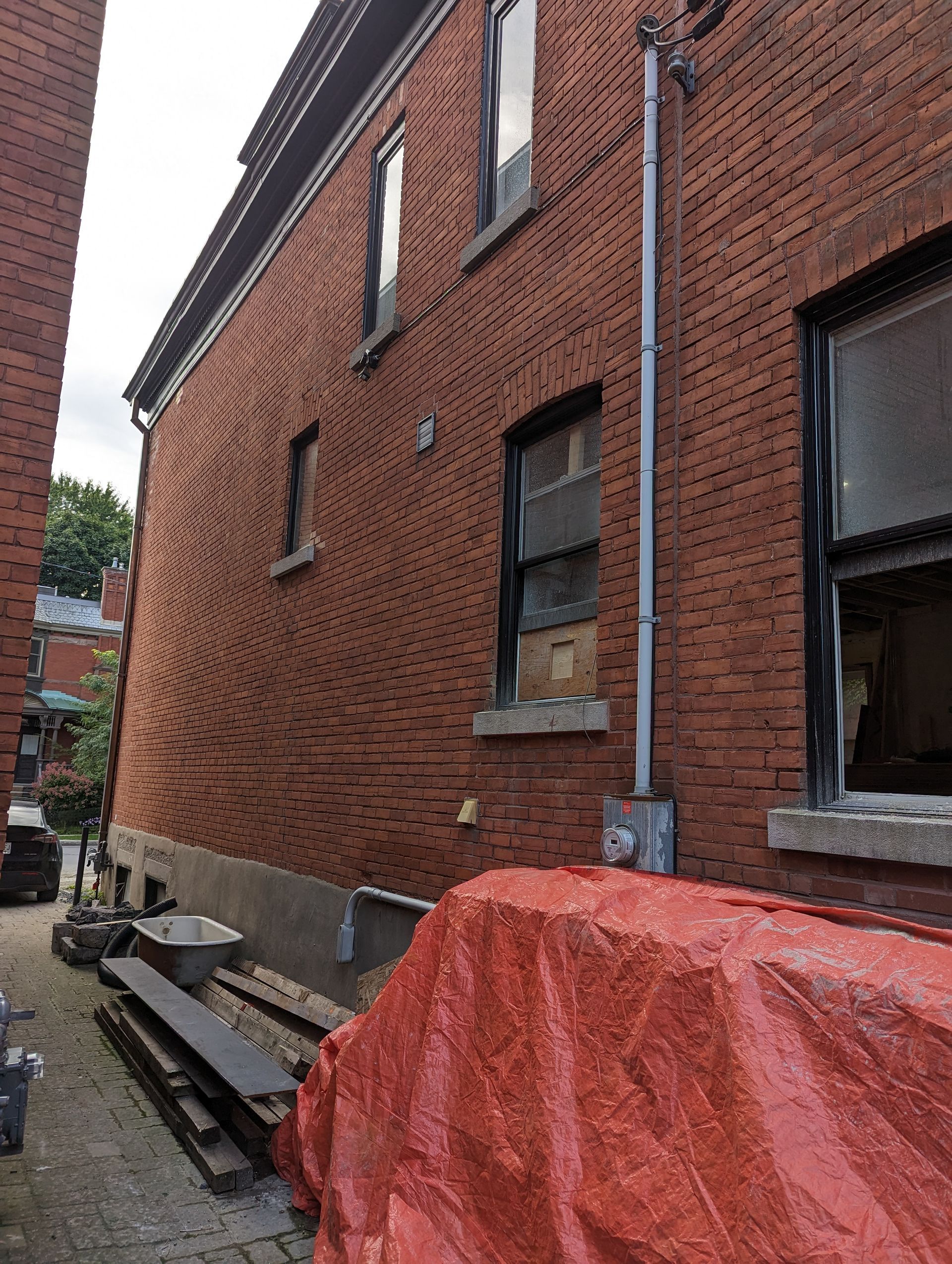 Façade d'un bâtiment en briques rouges avec fenêtres, compteur électrique et bâche rouge dans une ruelle.