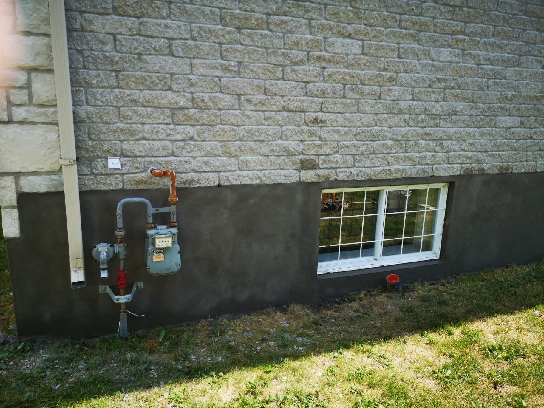 Vue extérieure d'un mur de maison en pierre avec une fenêtre de sous-sol, une descente de gouttière et un compteur de gaz