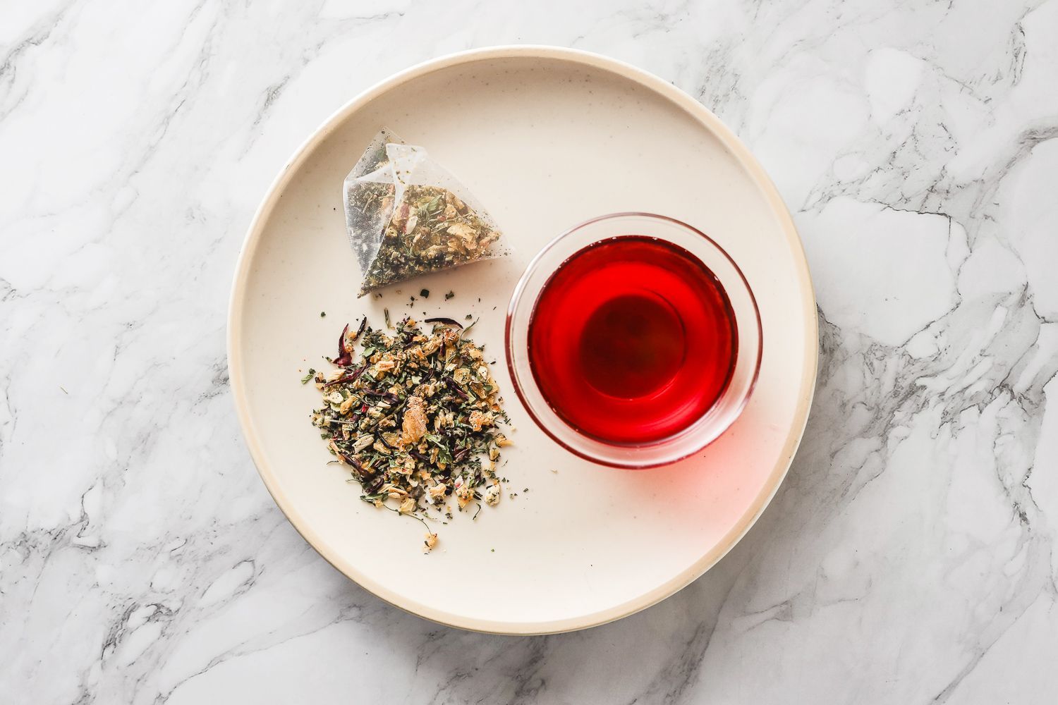 一個茶包、一些散裝茶葉和一小杯鮮紅色的液體放在白色盤子上,盤子靠在大理石檯面上。