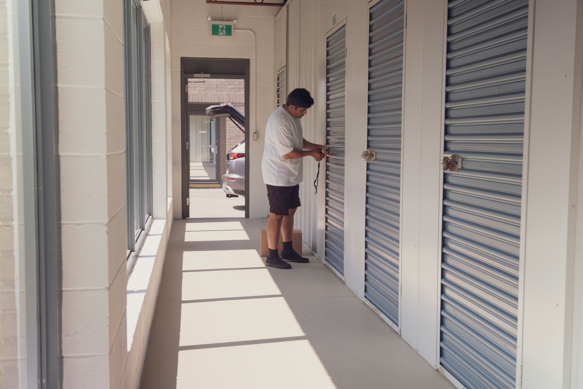 A man is standing in a hallway with blue storage units. He is working on a lock.