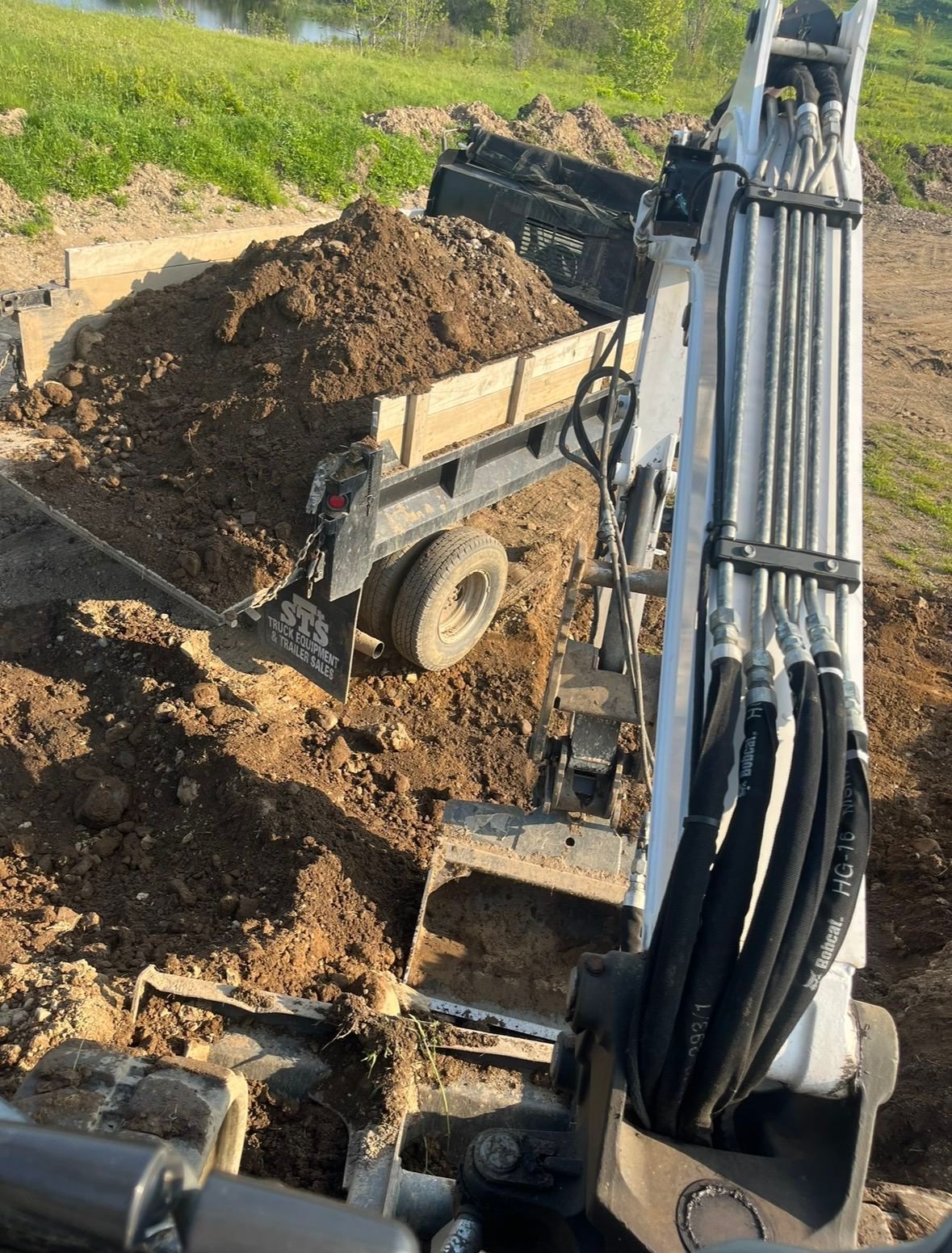 An excavator bucket sits in the foreground, ready to dump dirt into a nearby trailer on a grassy, outdoor work site.