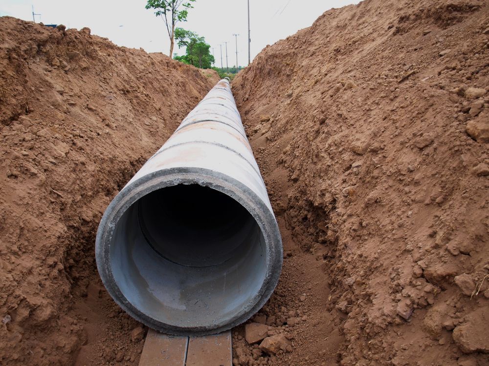 A large, circular concrete drainage pipe sits centered in a deep trench of reddish-brown dirt.