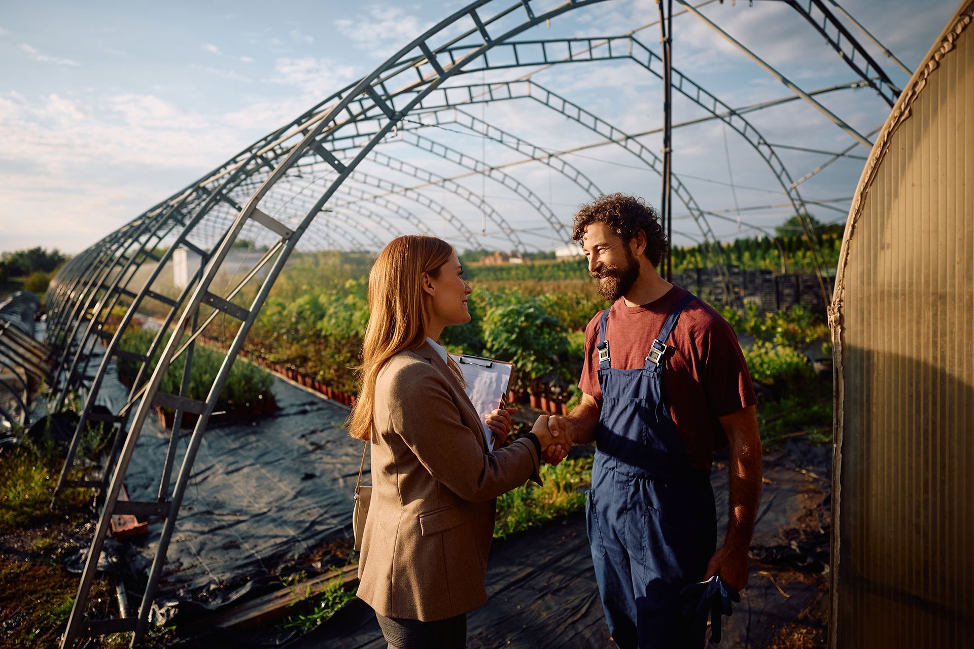 Woman and man shaking hands inside greenhouse, plants visible; outdoors.