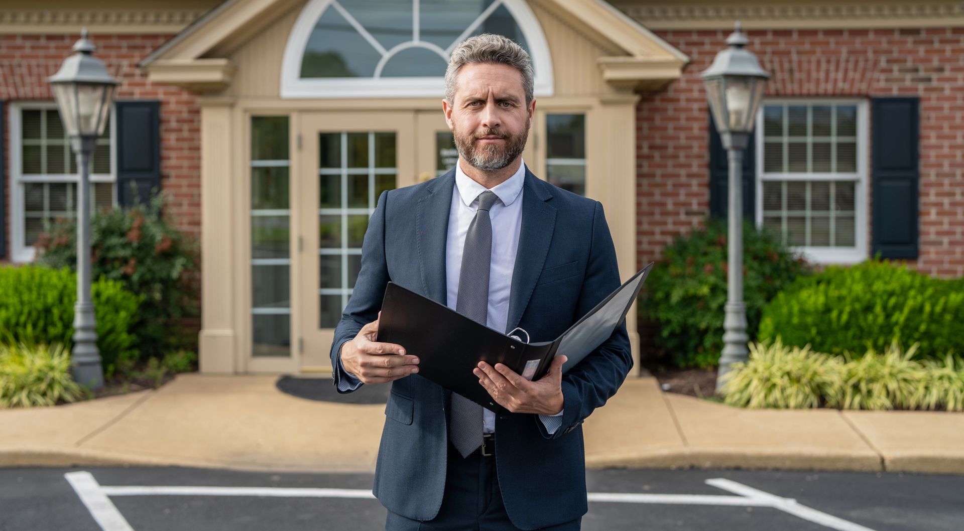 Renters insurance agent in a dark gray suit holding an open binder outside a brick rental property.