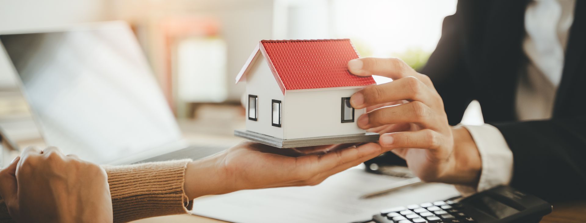 Hands exchanging a small house model over a desk with a laptop in the background.