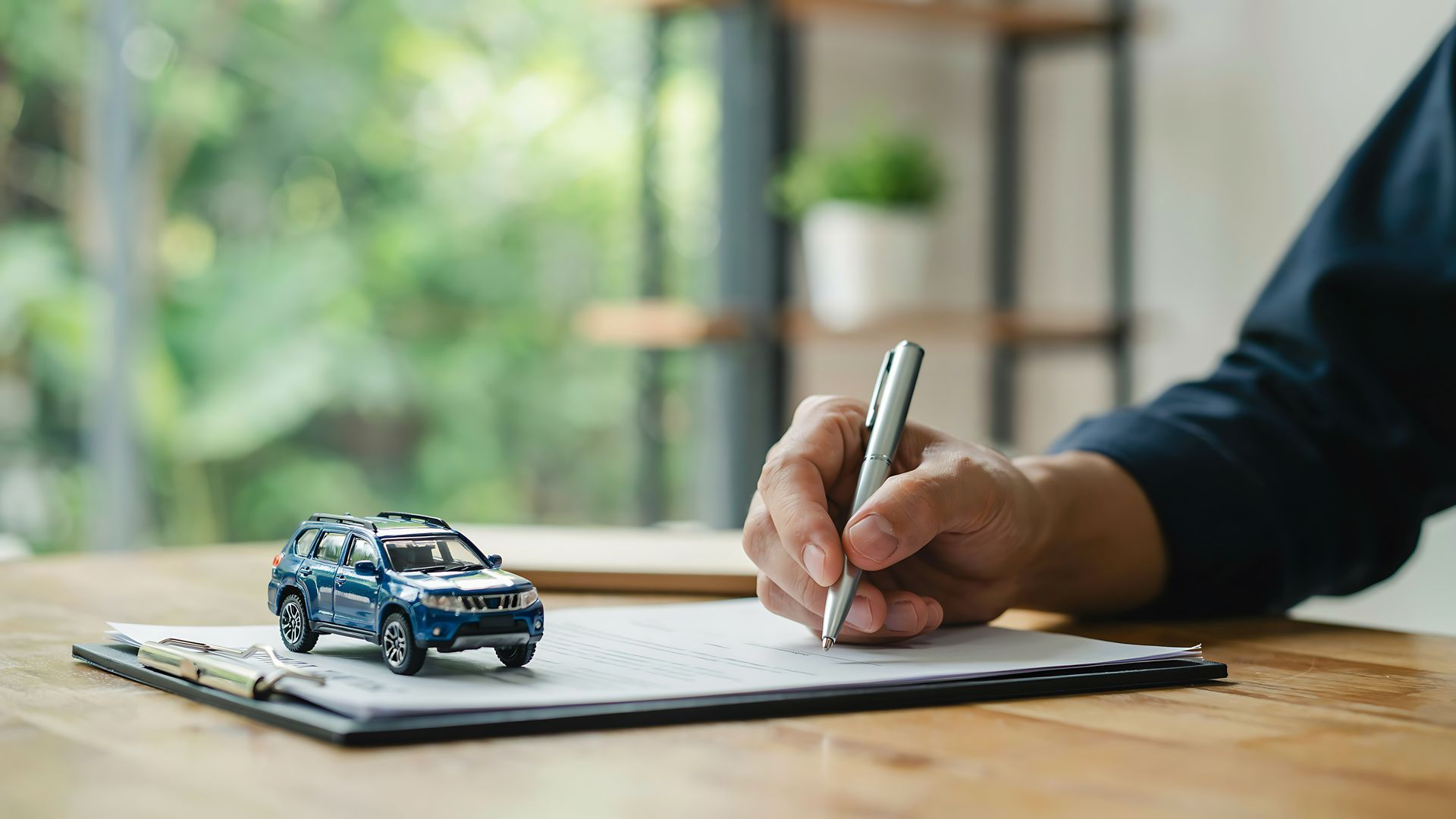 Insurance agent in a suit signing paperwork at a desk beside a model house and a set of house keys. Business owner signing a commercial auto insurance policy beside a blue SUV model and vehicle keys.
