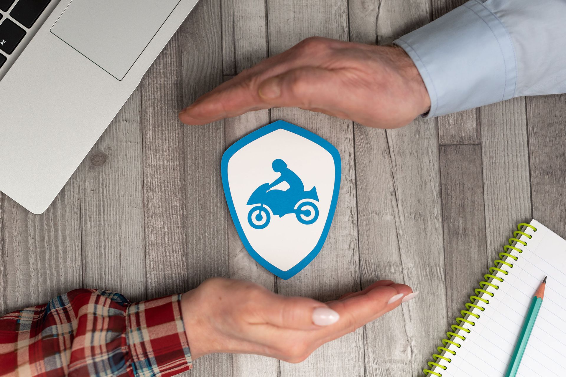 Hands protecting a blue motorcycle icon on a shield over a wooden surface, with a laptop and notepad nearby.