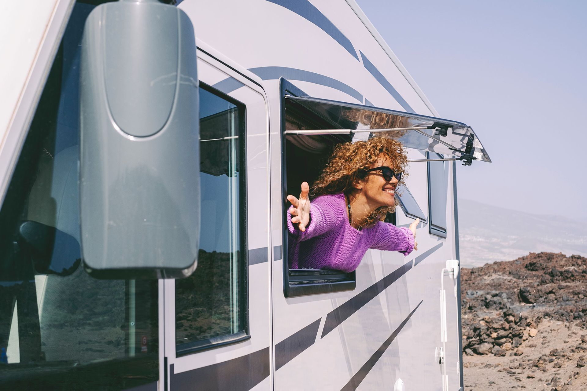 Woman leaning out RV window with arms outstretched, rocky landscape.