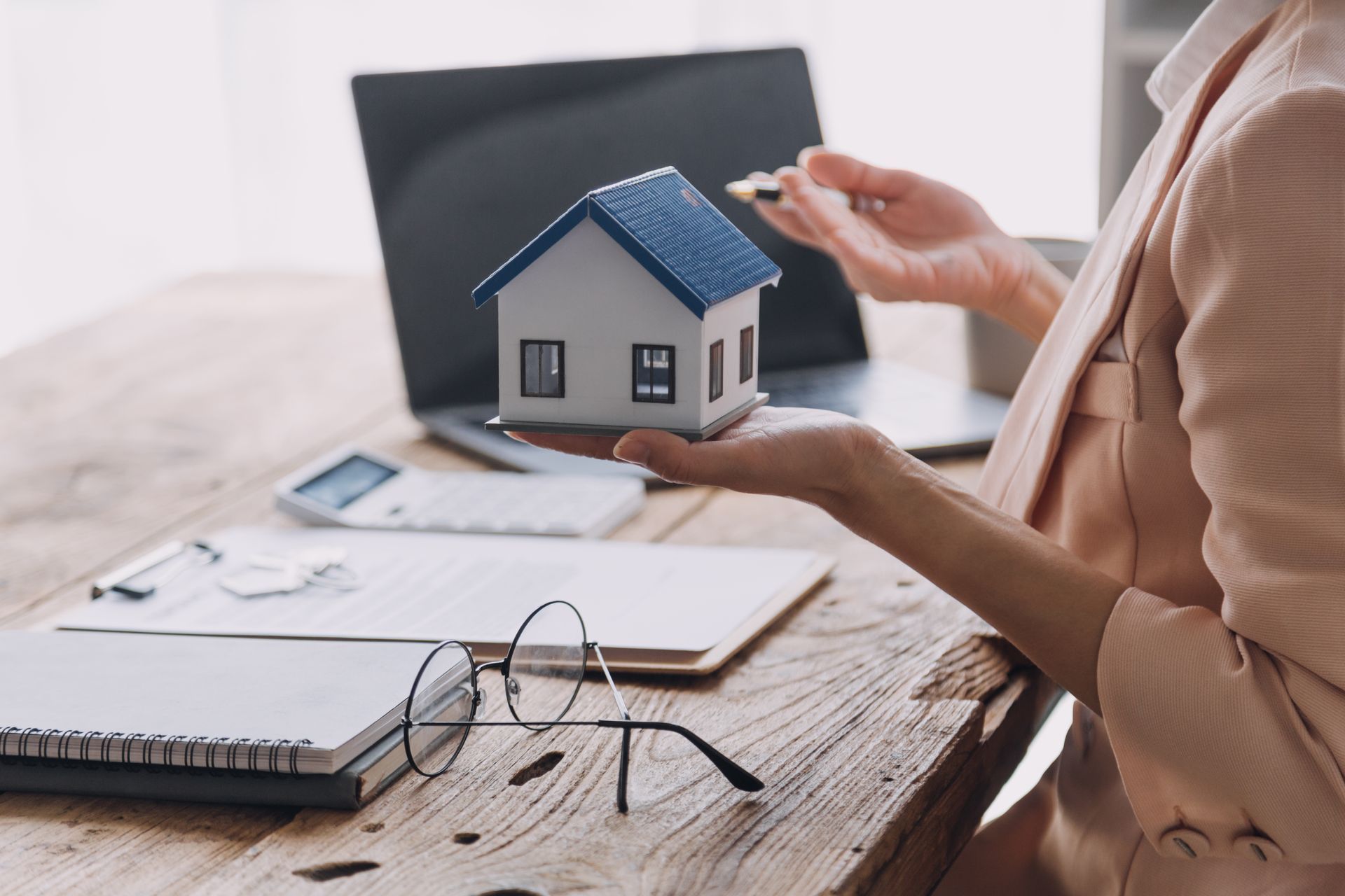 Person holding a small house model over a desk with laptop, papers, and glasses.