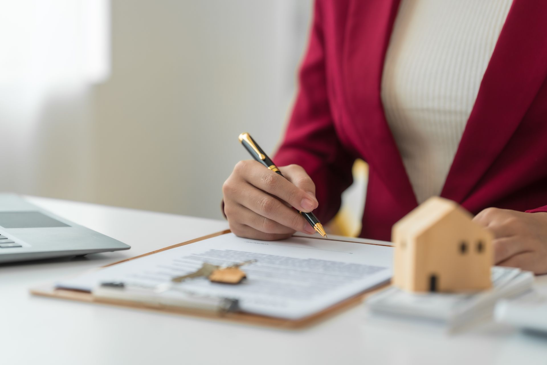 Person in red blazer writing on a document beside a laptop and small house model.