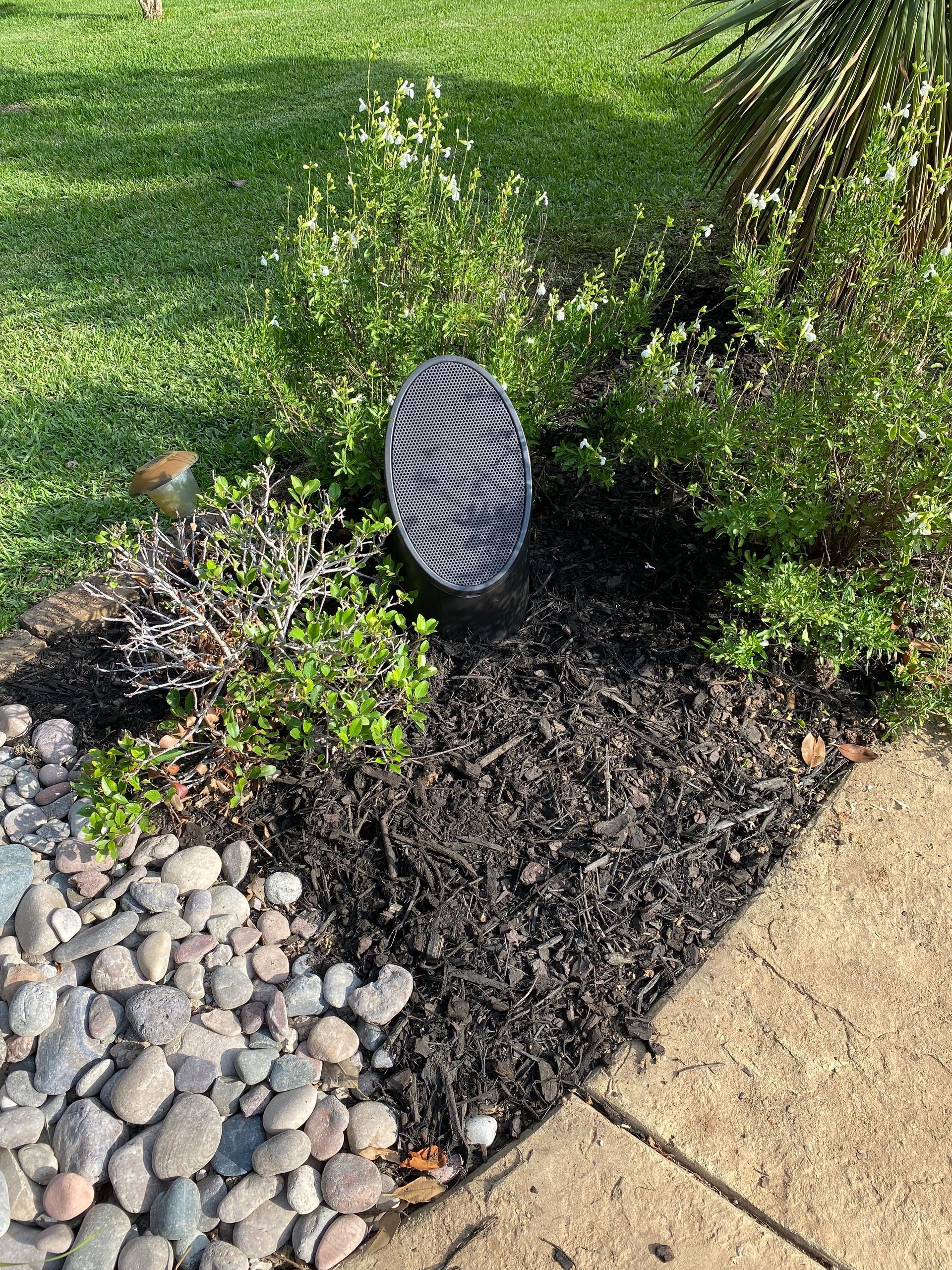 A speaker is sitting in the middle of a garden next to a sidewalk.