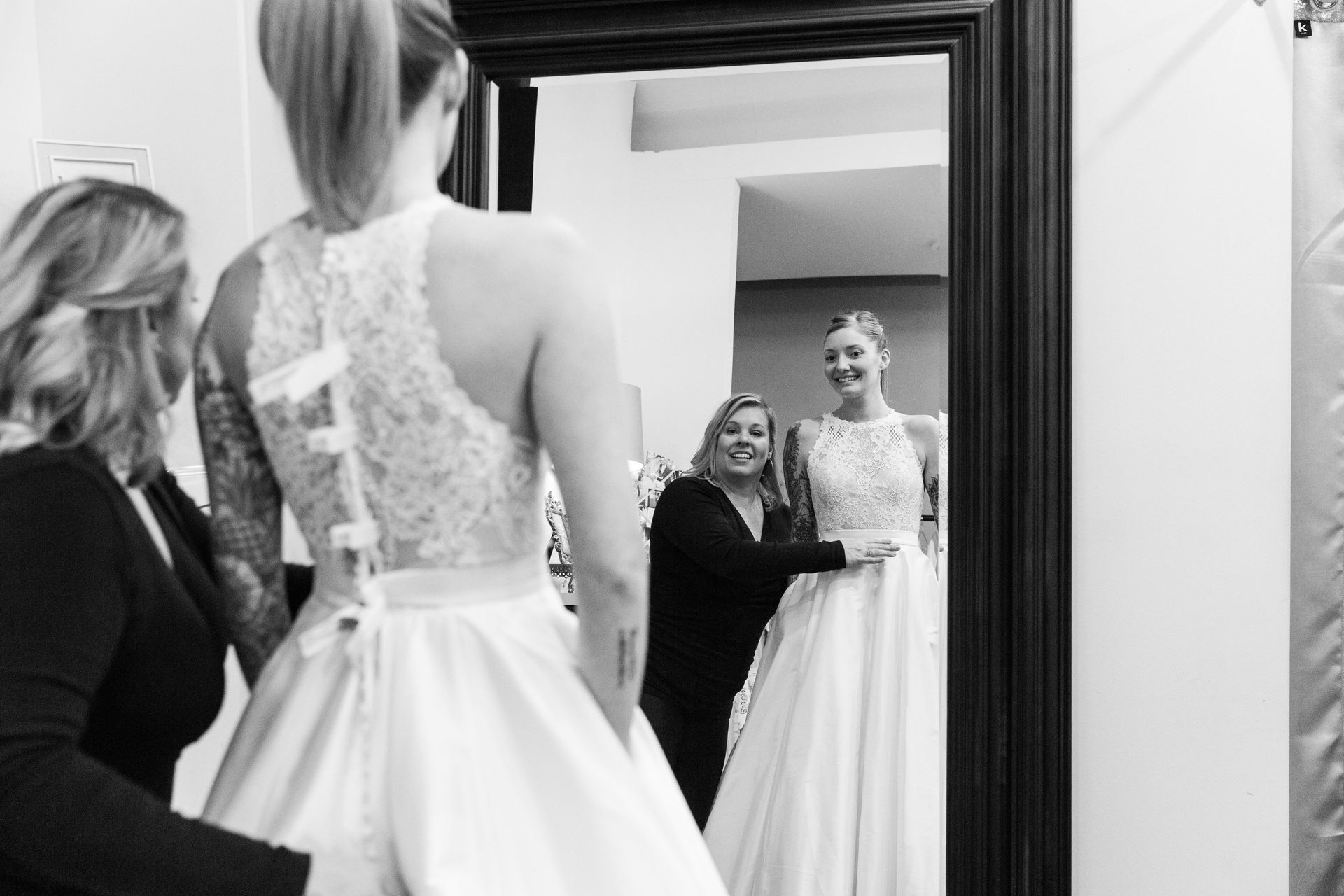Bride in a white gown looks at her reflection in a mirror while two women help her get ready.