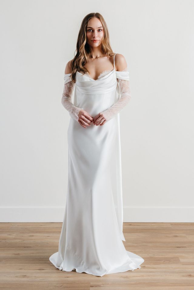 Woman in a white off-shoulder gown standing against a plain wall on a wooden floor