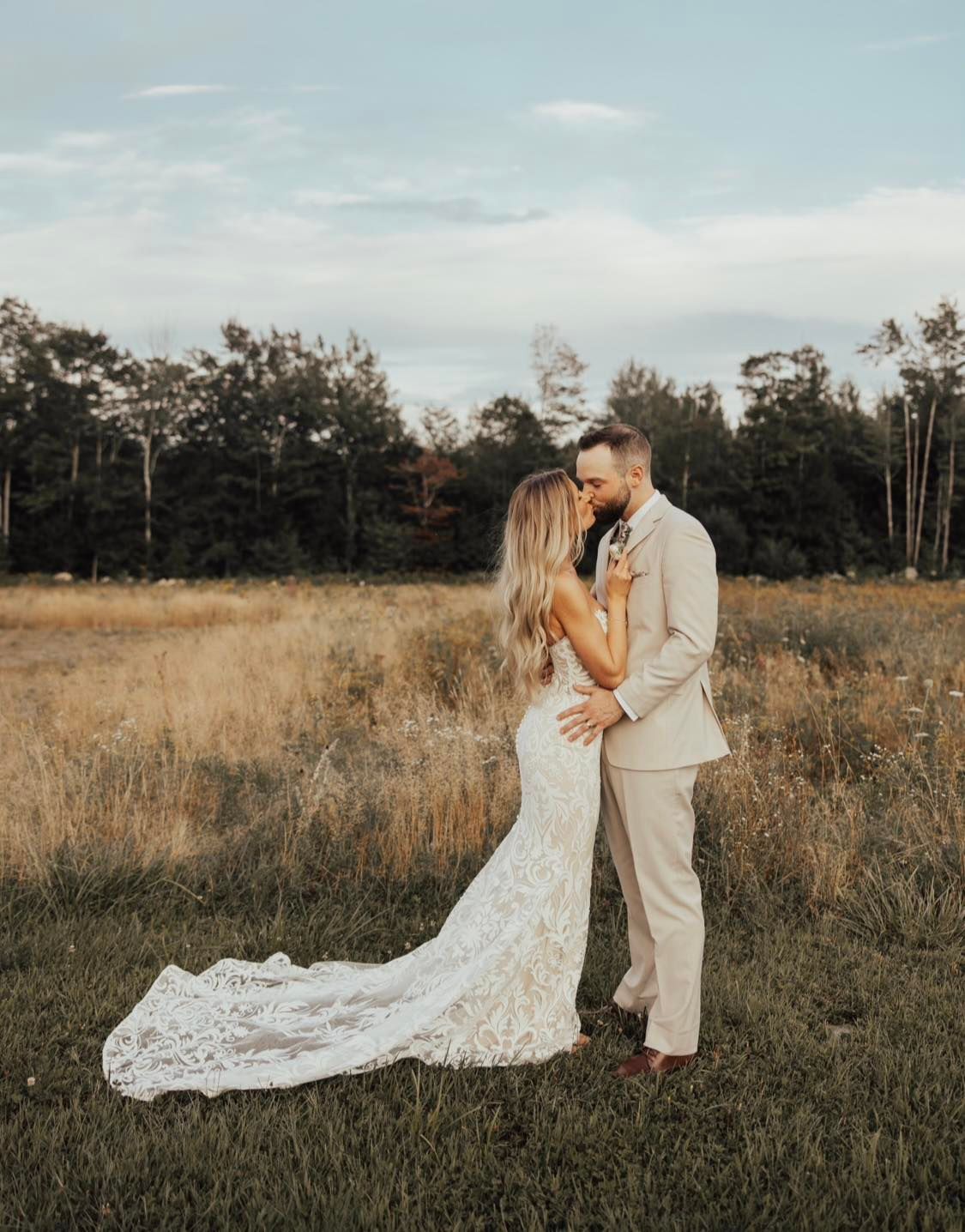 Couple embracing in a grassy field at sunset, with the bride’s lace train flowing behind them.