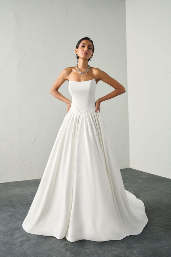 Woman in a strapless white gown posing in a minimalist studio with hands on hips