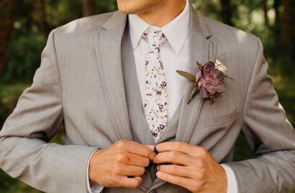 Man in a light gray suit buttoning his jacket, wearing a floral tie and boutonniere outdoors