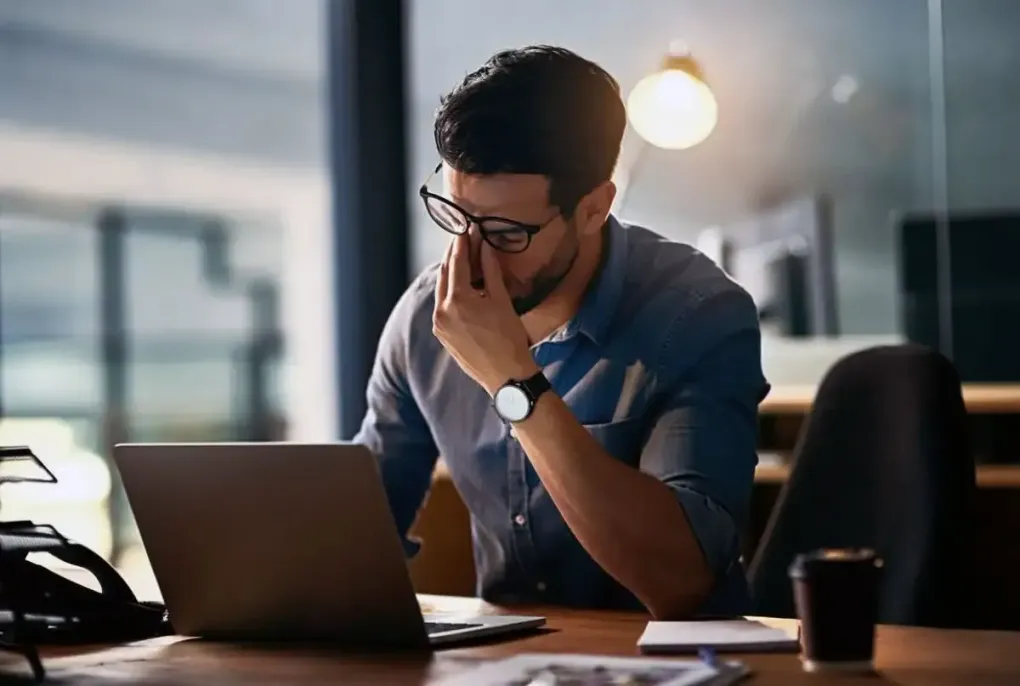 Man at a desk with a laptop, looking thoughtful in a dim office with a coffee cup nearby.