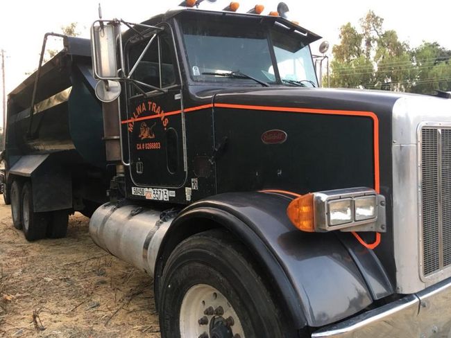 Black semi-truck with chrome grille parked on dirt road, carrying a load on the trailer