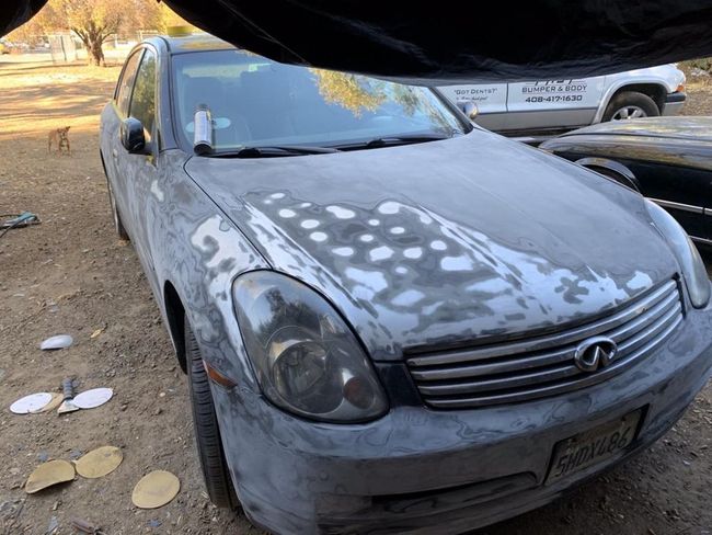 Gray car with white repair spots on the hood, parked outdoors under a canopy.
