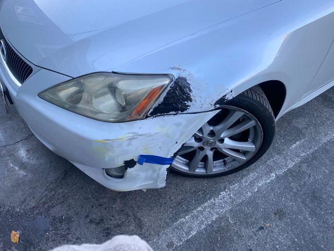 Damaged white car front bumper and headlight after collision, parked on pavement.