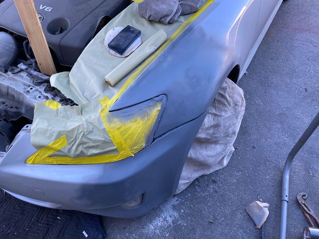 Grey car front bumper masked with tape and plastic, being prepped for painting or repair
