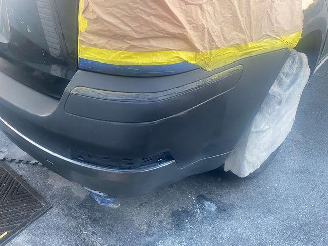 Rear bumper of a black car with scuff damage, partially covered with masking tape and plastic wrap.